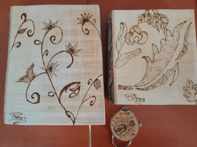 Dwa kawałki drewna z motywami roślinnymi wykonane techniką wypalania w drewnie. Na jednym ptaszek i kwiaty, na drugim liście i kwiaty. Obok breloczek z gitarą.