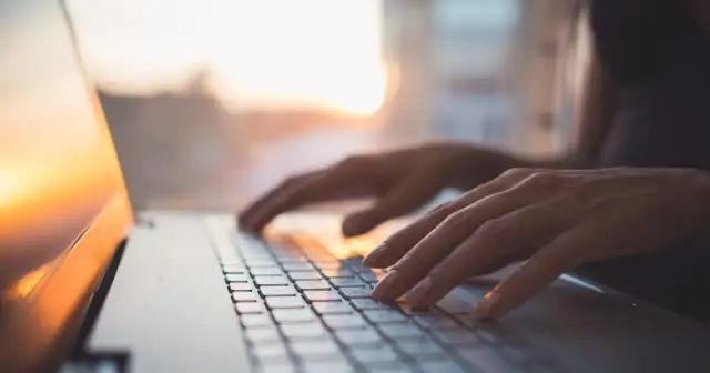 Dłonie piszą na klawiaturze laptopa z systemem MX Linux, w tle zachodzące słońce.