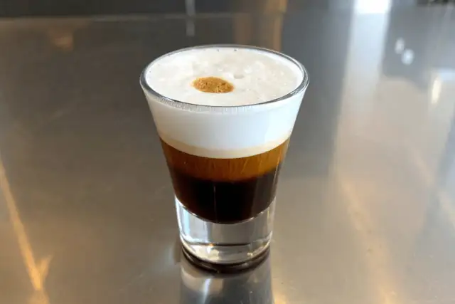 Kawa caffe coretto w szklance, z warstwami ciemnego espresso i białej pianki, ozdobiona złotą kropką.