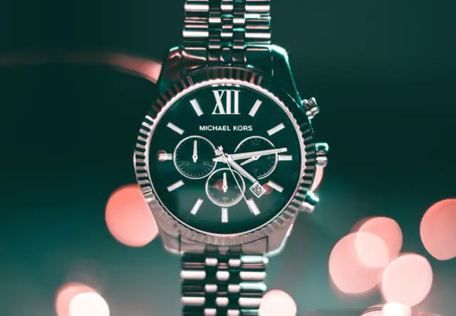Elegancki zegarek Michael Kors z ciemną tarczą i srebrną bransoletą, na tle rozmytych świateł.