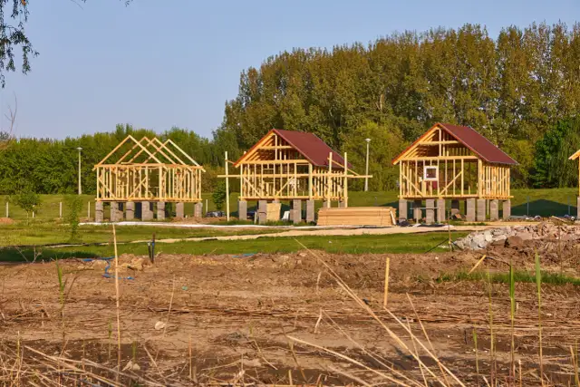 Trzy drewniane szkielety budynków gospodarczych na betonowych fundamentach, w tle drzewa.