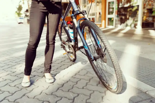 Osoba w czarnych spodniach i balerinkach stoi obok niebieskiego roweru na chodniku.