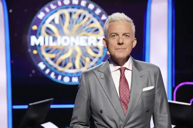 Hubert Urbański w garniturze, na tle koła z napisem "Milionerzy". Co dajemy komuś na drogę milionerzy? Wiedzę i odwagę!