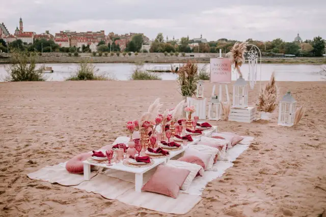 Elegancki wieczór panieński w plenerze nad rzeką. Stół z różowymi kieliszkami, serwetkami i kwiatami. W tle miasto.