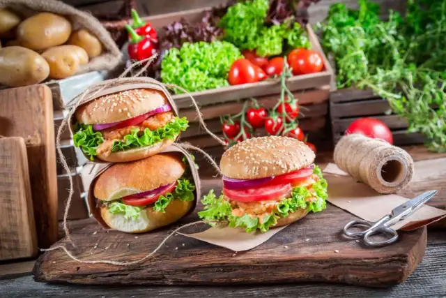 Dwa apetyczne burgery z sałatą, pomidorem i cebulą na drewnianej desce, w tle warzywa.