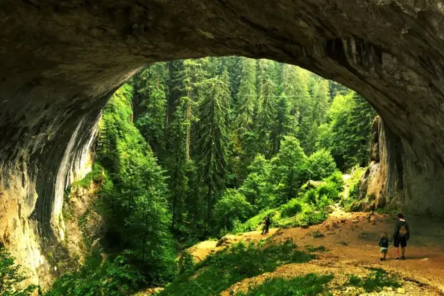 Wspaniałe skalne okno w górach Rodopy otwiera widok na bujny, zielony las. Ludzie spacerują ścieżką.