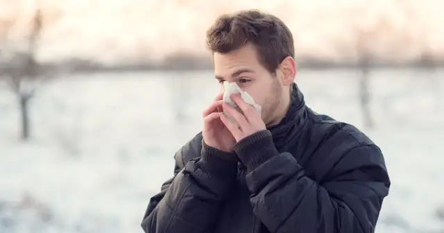 Młody mężczyzna w zimowej kurtce chowa twarz w chusteczkę, cierpiąc na przewlekły mokry kaszel.