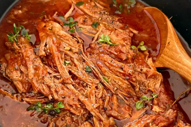 Miękka, szarpana wołowina w sosie pomidorowym, ozdobiona ziołami, obok drewnianej łyżki.