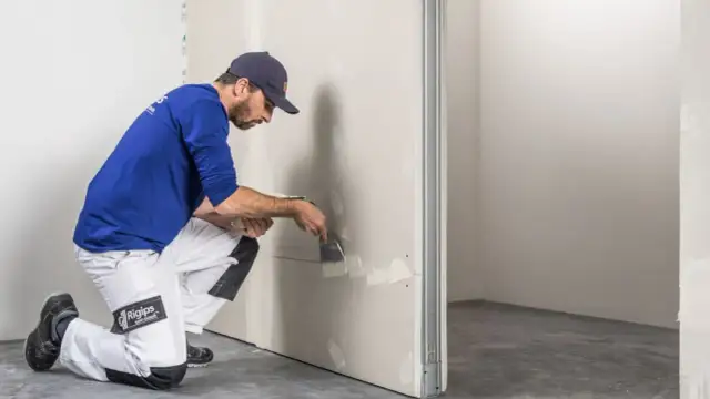 Murarz w niebieskiej koszulce i białych spodniach nakłada gładź na płytę gipsowo-kartonową.