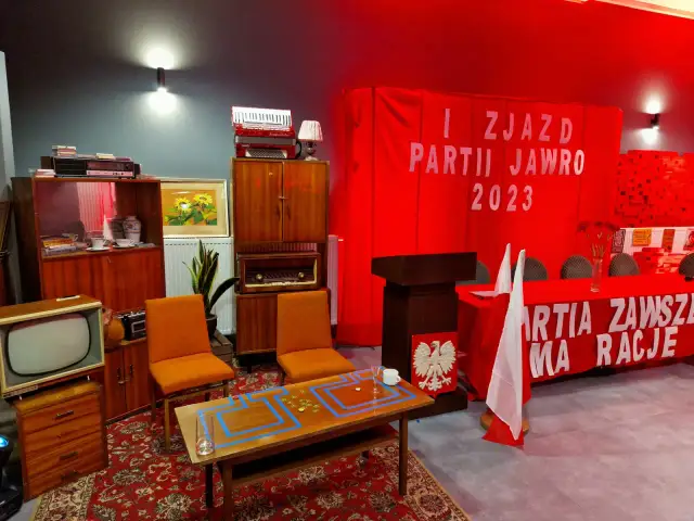 Wystrój w stylu PRL: meble, radio, akordeon, czerwony transparent z napisem "PARTIA ZAWSZE MA RACJE".