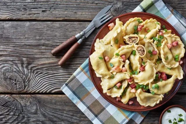 Pyszne pierogi z farszem na pierogi z mięsem mielonym, posypane szczypiorkiem i skwarkami. Obok widelec i nóż.
