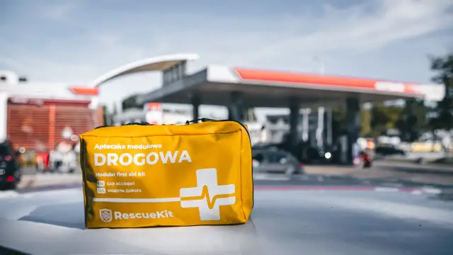 Żółta apteczka samochodowa z napisem "DROGOWA" na tle stacji benzynowej.
