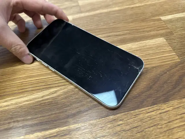Dłoń trzyma telefon z pękniętym szkłem hartowanym. Ekran pokryty jest siecią drobnych pęknięć, szczególnie widocznych w prawym dolnym rogu.