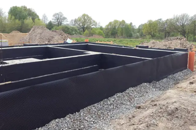 Fundament domu z czarną izolacją przeciwwilgociową i drenażową, otoczony żwirem.