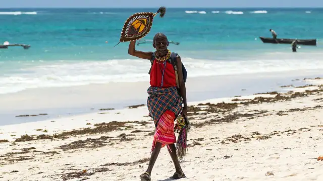Mężczyzna w tradycyjnym stroju na plaży w Kenii, trzymający ozdobną tarczę. Może to symbolizować bogactwo kulturowe, które jest cenniejsze niż kenijska waluta.