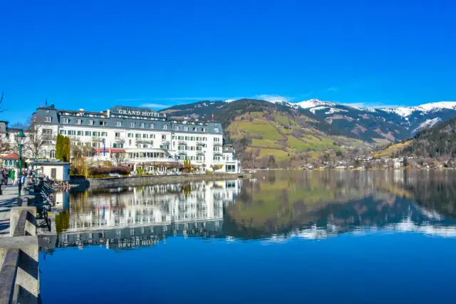Grand Hotel nad jeziorem w Zell am See, z odbiciem w spokojnej wodzie i ośnieżonymi górami w tle.