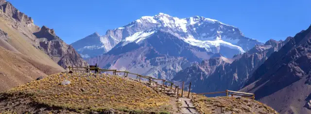 Majestatyczny Aconcagua szczyt w Andach, pokryty śniegiem, góruje nad suchym, skalistym krajobrazem. Ścieżka z drewnianą balustradą prowadzi do punktu widokowego.
