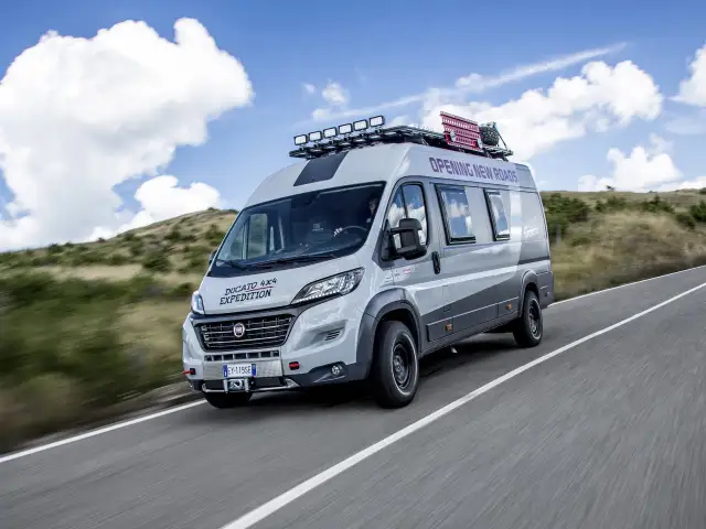 Szary kamper Ducato 4x4 Expedition z bagażnikiem dachowym i dodatkowym oświetleniem jedzie po drodze wśród wzgórz.