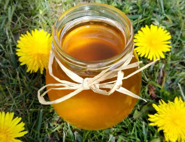 Słoik ze złotym syropem z mniszka, ozdobiony słomkową wstążką, otoczony polnymi kwiatami. Idealny syrop z mniszka przepis na wiosenne przetwory.