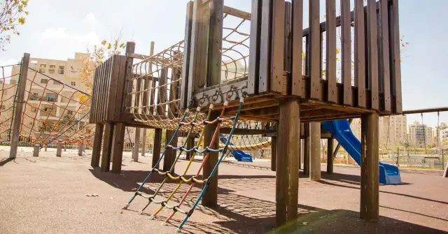 Drewniany plac zabaw z linami, zjeżdżalnią i siatkami w tle.