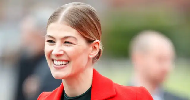 Uśmiechnięta Rosamund Pike, znana z wielu filmów i seriali, w czerwonej marynarce.