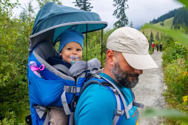 Wesoły maluch w nosidle na plecach taty podczas wyprawy w góry. Idealny pomysł, gdzie w góry z dzieckiem 2 letnim.