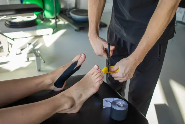 Fizjoterapeuta zakłada czarny plaster na stopę pacjenta, obok leży rolka plastra.