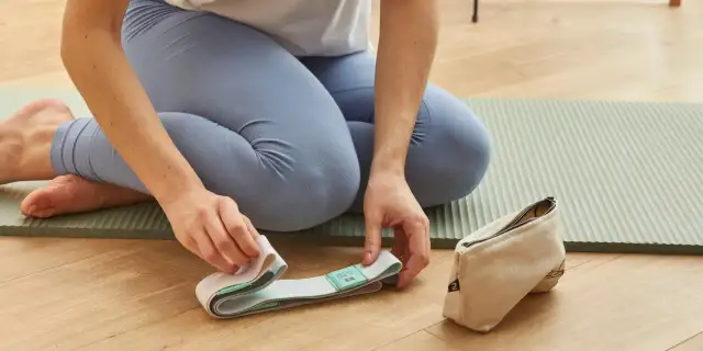 Kobieta w legginsach przygotowuje pasek do jogi na macie. Obok leży woreczek.