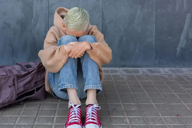 Młoda osoba w beżowej bluzie z kapturem i jeansach siedzi skulona na chodniku obok plecaka.