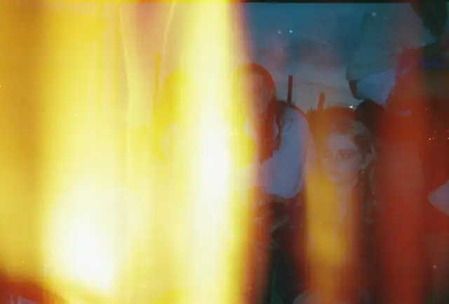 Jasne, oto tekst alternatywny:

Zdjęcie z aparatu jednorazowego z rozmytymi postaciami i jasnym, żółtym światłem.