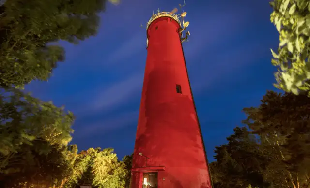 Czerwona latarnia morska w Krynicy Morskiej nocą, otoczona drzewami.