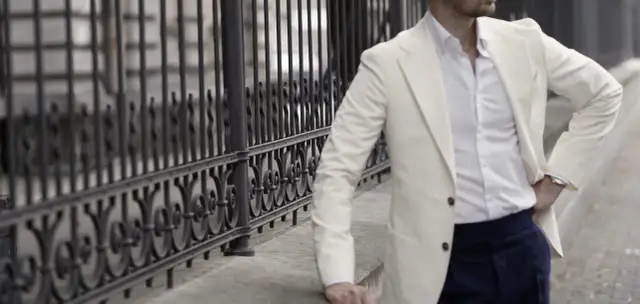 Mężczyzna w kremowej marynarce i białej koszuli opiera się o kamienny murek przy ozdobnej balustradzie.