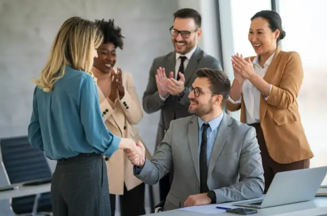 Zespół gratuluje pracownicy, ściskając jej dłoń. Wszyscy uśmiechnięci, to wyraz podziękowania za zaangażowanie.