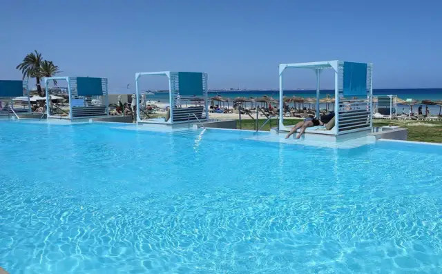 Basen z turkusową wodą, leżaki z baldachimami i plaża w tle.