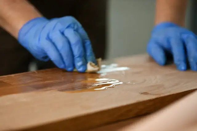 Ręce w niebieskich rękawiczkach nakładają środek na drewniany blat.