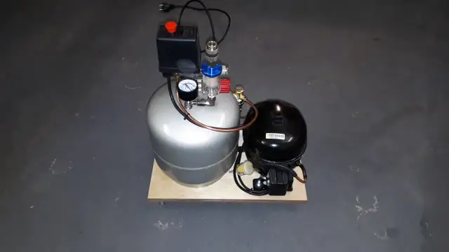 Srebrny kompresor z czarnym agregatem i manometrem na drewnianej podstawie.