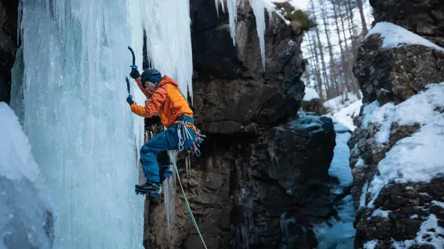 Wspinaczka górska po lodospadzie. Alpinista w pomarańczowej kurtce i niebieskich spodniach wspina się po zamarzniętej ścianie, używając czekana i raków.