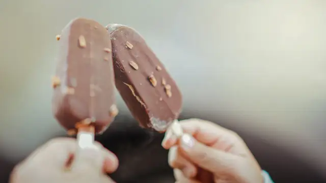 Dwie ręce trzymają czekoladowe lody na patyku z orzechami. Idealne na lato, wegańskie lody gdzie kupić?