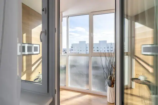 Zabudowany balkon z widokiem na budynki, ozdobiony wazonem z gałązkami.