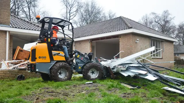 Pomarańczowa koparko-ładowarka z operatorem w kasku i kamizelce usuwa gruz z posesji.