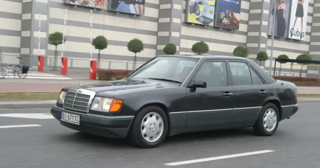 Ciemnoszary Mercedes W124 jedzie ulicą przed budynkiem z banerami.