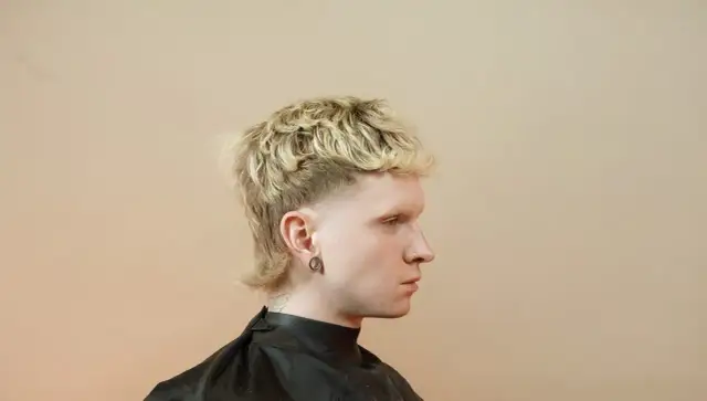 Młody mężczyzna z blond, kręconymi włosami i charakterystyczną fryzurą mullet.