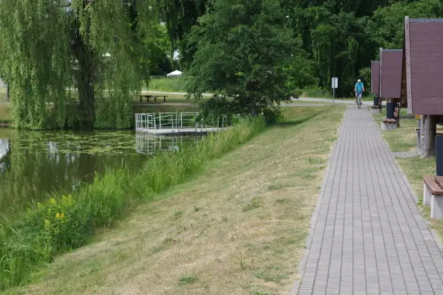 Ścieżka z kostki brukowej nad jeziorem z pomostem, w tle osoba na rowerze i domki.