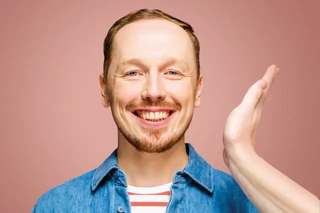 Uśmiechnięty mężczyzna w dżinsowej koszuli, jakby właśnie usłyszał świetny żart na występie syrek dąbrowski stand up.