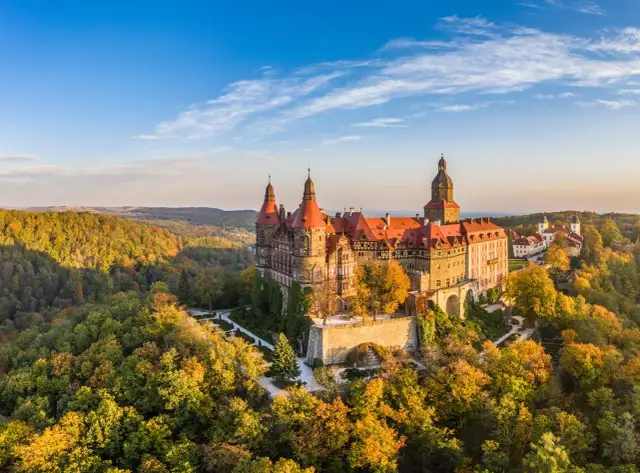 Zamek Książ w malowniczej kotlinie kłodzkiej, otoczony jesiennymi barwami lasów, prezentuje się majestatycznie na tle błękitnego nieba.