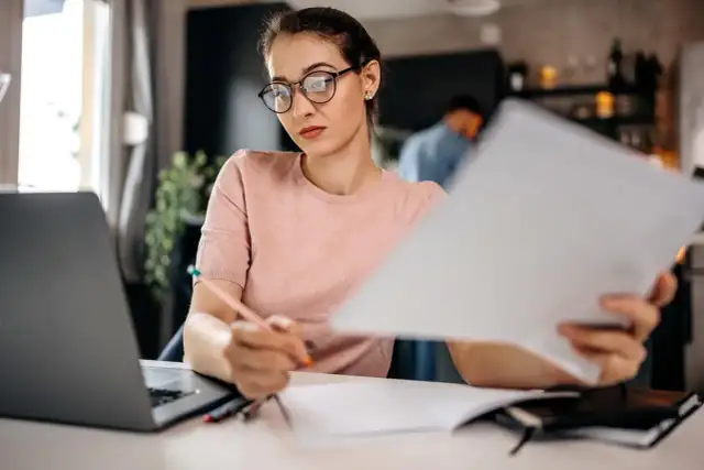 Kobieta w okularach pracuje przy biurku z laptopem i dokumentami.