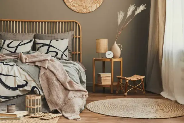 Dormitorio con cama de ratán, ropa de cama beige y gris, manta rosa, mesita auxiliar de madera y alfombra redonda.