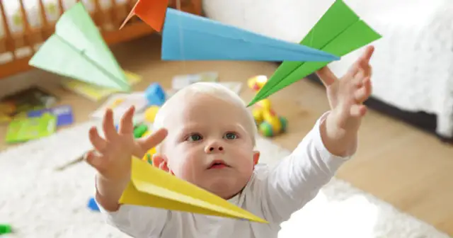 Niemowlę z wyciągniętymi rączkami próbuje złapać kolorowe papierowe samolociki.