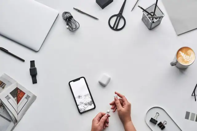 Dłonie trzymające białe słuchawki AirPods obok smartfona i etui.