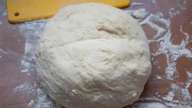 Surowe ciasto na pizzę na drewnianym blacie, posypane mąką. Obok żółta łopatka.